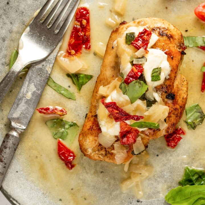 A grey plate with a chicken breast on it. The chicken breast is topped with goat cheese, sun-dried tomatoes, and basil.