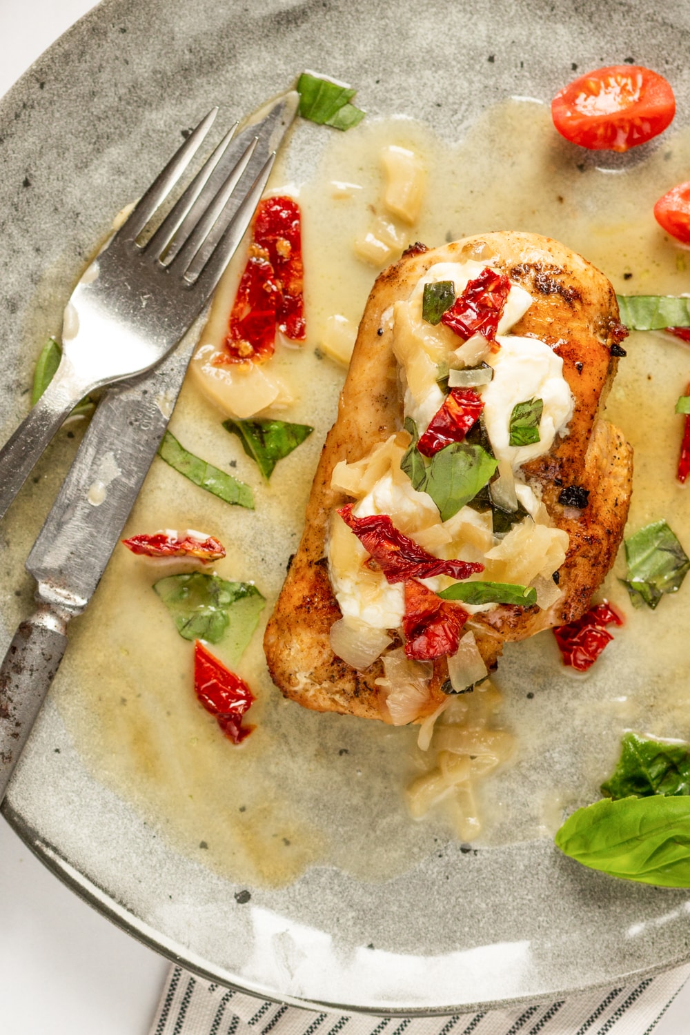 A grey plate with a chicken breast on it. The chicken breast is topped with goat cheese, sun-dried tomatoes, and basil.