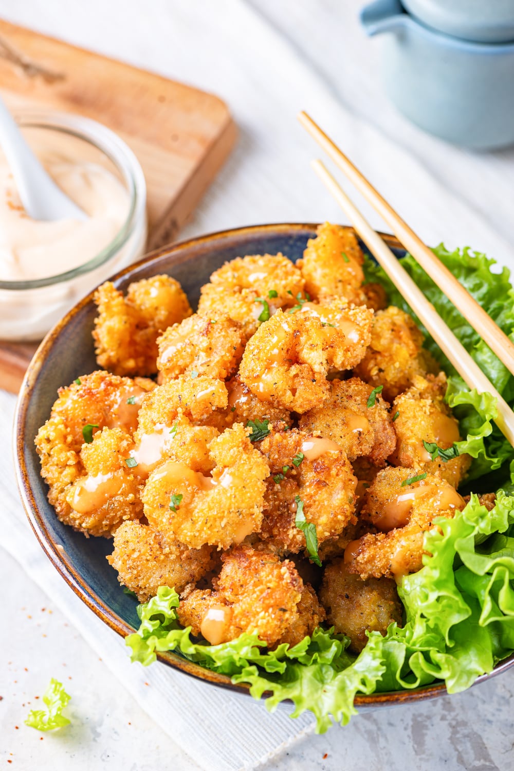 A blue bowl filled with bang bang shrimp. There is a lettuce garnish around the outer right rim of the bowl with chopsticks laid on top. A small glass jar of bang bang sauce is on a wooden cutting board to the left behind the bowl of shrimp.