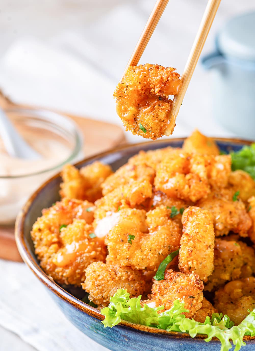 A blue bowl filled with bang bang shrimp with a lettuce garnish at the front rim of the bowl. A pair of chopsticks is hovering over the bowl with a piece of shrimp between the chopsticks. A small glass jar of bang bang sauce is on a wooden cutting board at the back left behind the bowl. Everything is on a white tablecloth on a white counter.