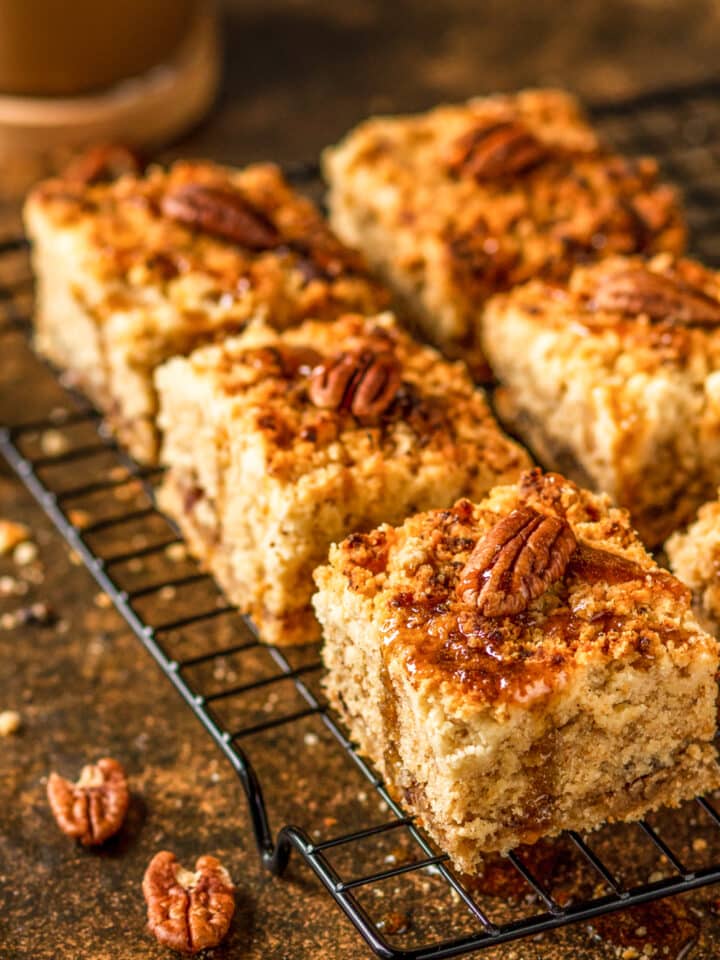 Three pieces of gluten free coffee cake in a row with two pieces to the side in a blurred view. There is one pecan on each piece of coffee cake and the coffee cake is on a wire rack on top of stone.