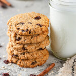 A stack of five vegan oatmeal raisin cookies with a tall glass jar of milk next to it on the right. There is a cinnamon stick in front of the cookies and milk with a few oats around them. Everything is on a grey counter.