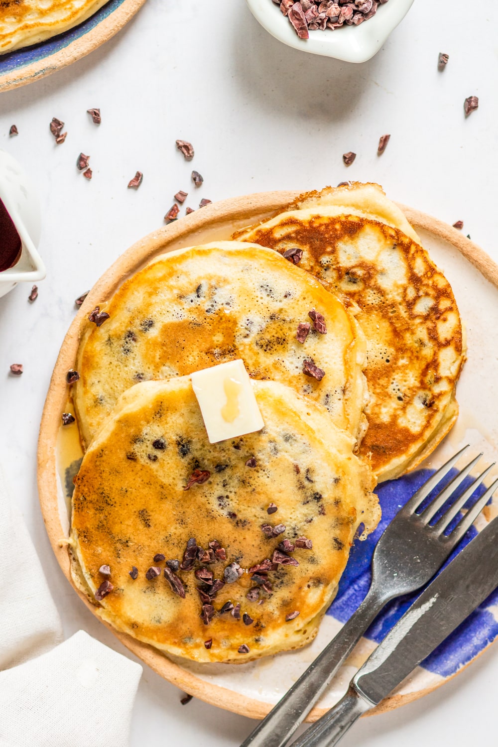 An overview of three chocolate chip pancakes overlapping one another on a white plate. There is a square slab of butter on the edge of the first pancake with a fork and knife at the bottom of the plate.