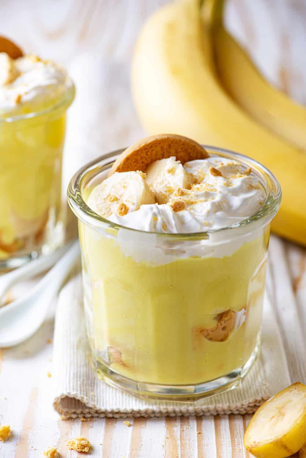 A glass cup filled with vegan banana pudding with vegan whipped cream, slices of bananas, and a vanilla cookie on top. Two bananas are behind the glass cup and the cup is on a cloth on a wooden table.