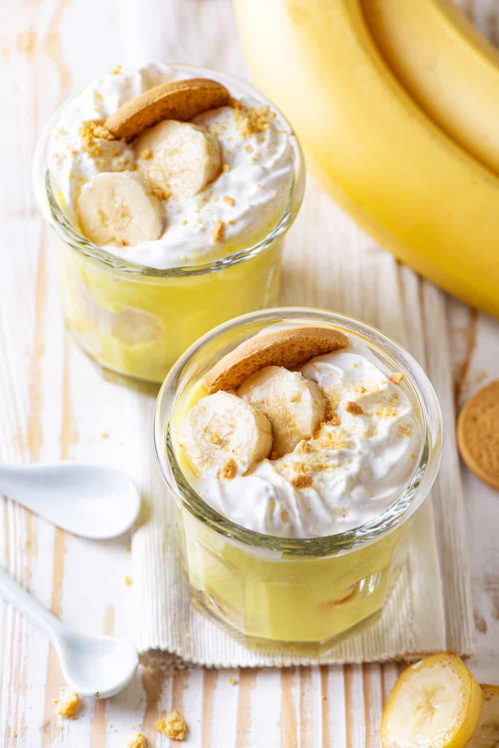 Two glass cups, one in front of the other, filled with vegan banana pudding. Each cup has vegan whipped cream, sliced bananas, and a vanilla cookie on top. The front cup is on a cloth and everything is on a wood table.