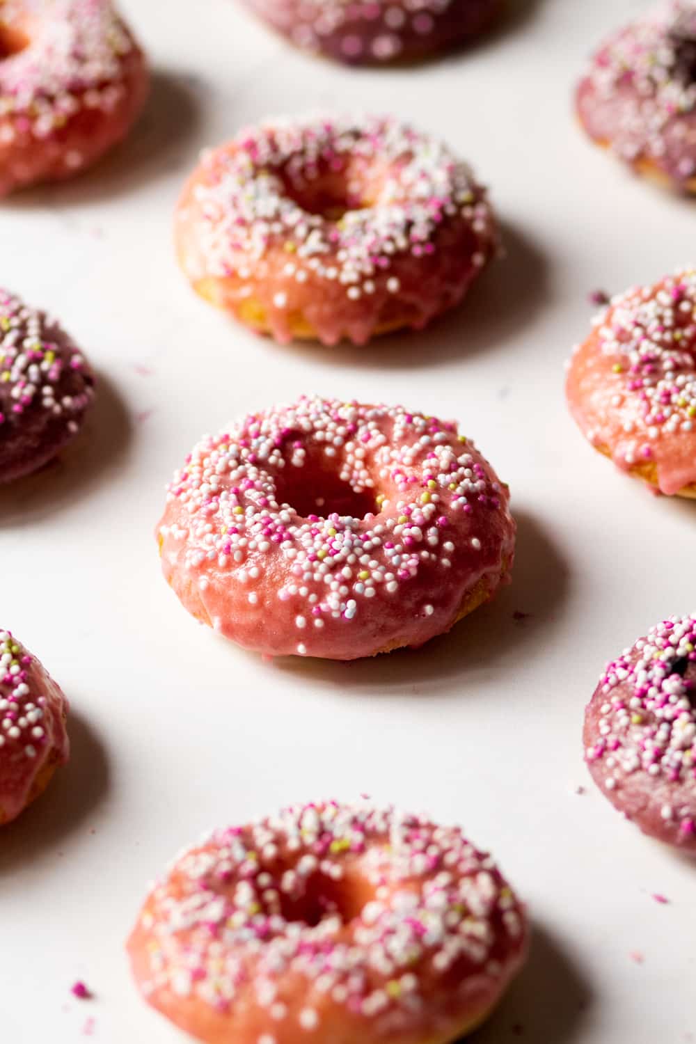 A row of three vegan donuts with pink glaze and white, pink, and yellow ball sprinkles on top. The donuts are on a white counter with parts of donuts surrounding them.