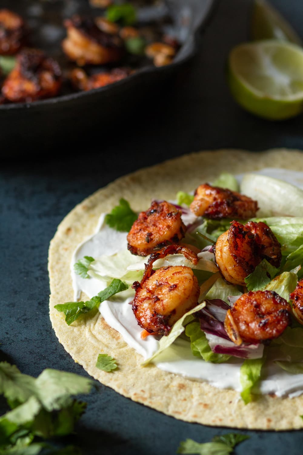 A tortilla shell with sour cream spread on it and a few pieces of blackened shrimp and some shredded lettuce on top. The tortilla shell is laying flat on a blue counter and a skillet filled with blackened shrimp is set behind the shell.