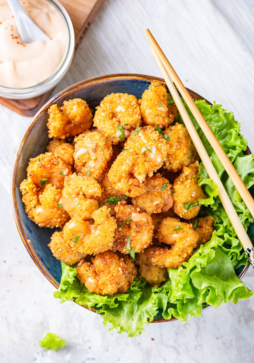 An overhead view of a blue bowl filled with bang bang shrimp. A lettuce garnish is on the rim of the right half of the bowl with chopsticks laid on top. A glass jar of bang bang sauce is to the back left of the bowl of shrimp. Everything is on a white counter top.