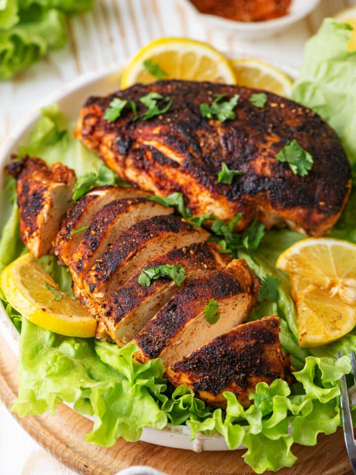 Two pieces of blackened chicken on a white plate with leafy green lettuce underneath the chicken. The piece of chicken at the front of the plate is cut into slices and the other piece of chicken at the back of the plate is whole. There are four slices of lemon on the side and back of the chicken. A white bowl of seasonings is behind the plate of chicken. The plate is on a wooden sauce and everything is on a wood table.