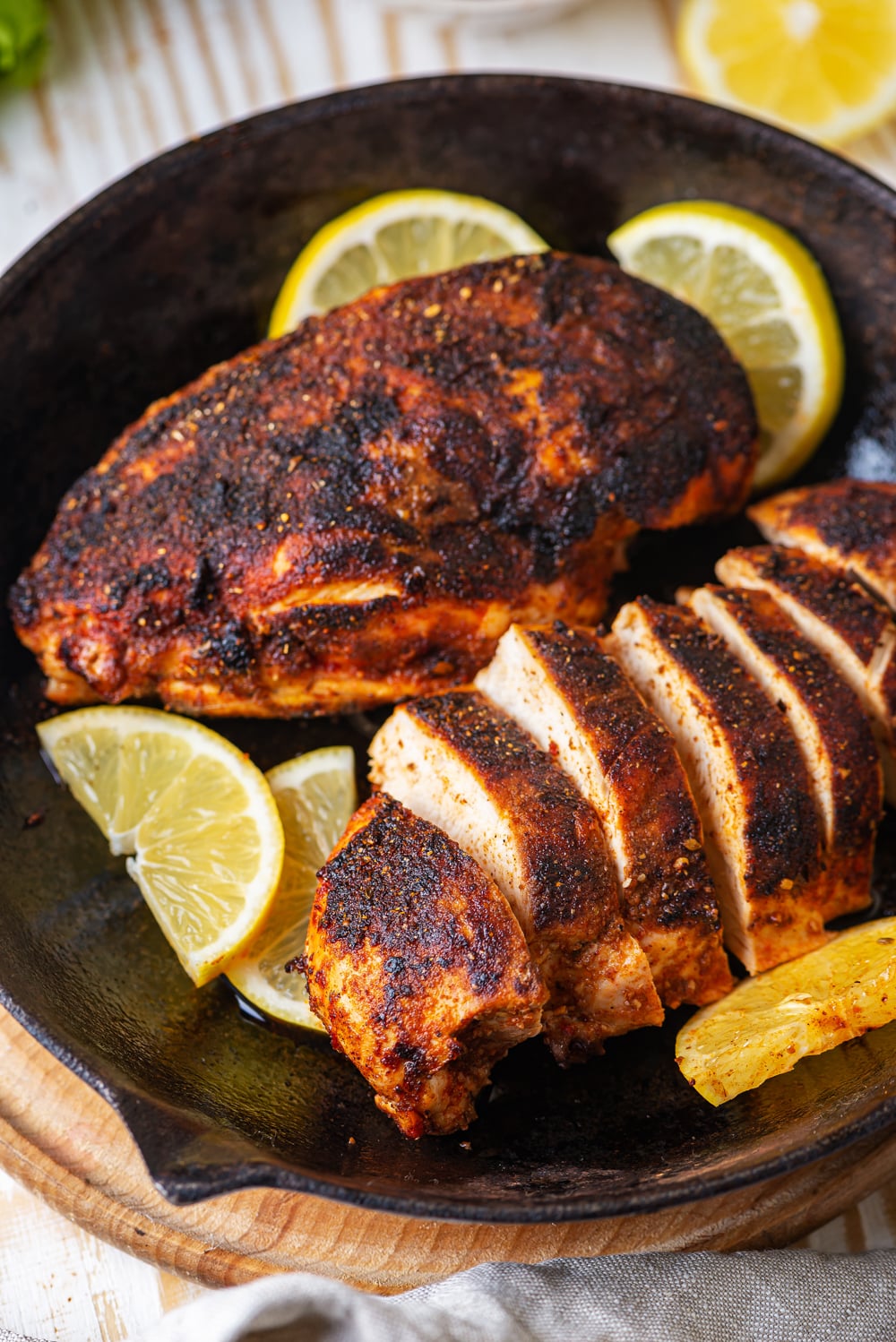 Two pieces of cooked blackened chicken in a skillet. The piece of chicken in the back of the skillet is whole and the front piece is cut into strips. There are slices of lemon around the chicken. The skillet is on a wooden saucer on a wood table.