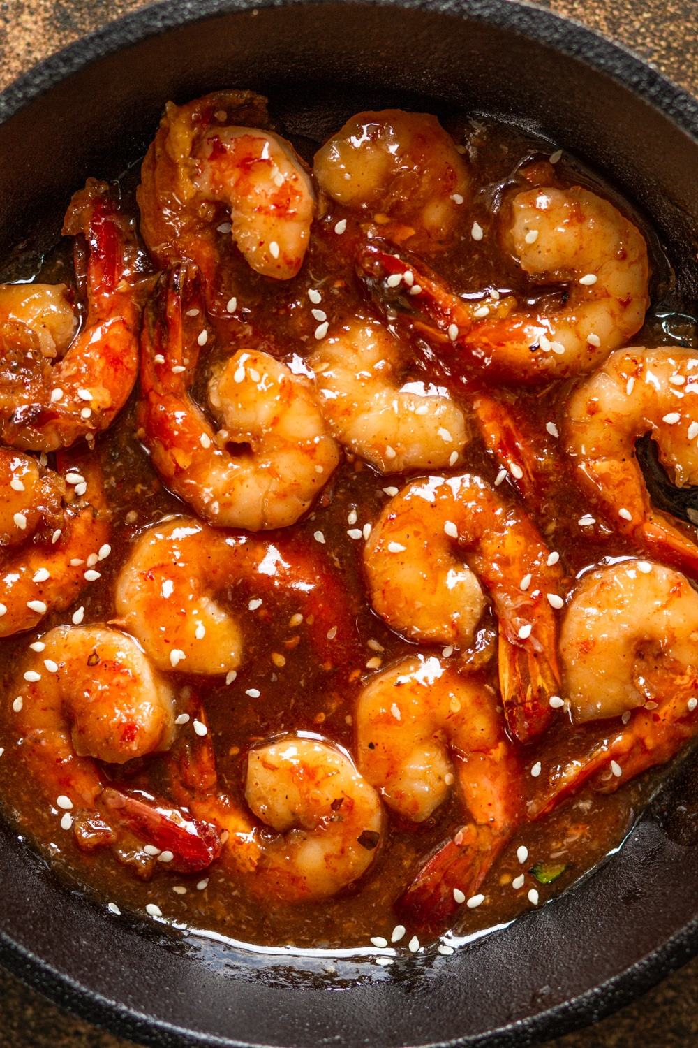 A close up overhead view of several pieces of shrimp in a skillet covered in firecracker sauce. There are sesame seeds scattered on the shrimp and in the sauce.
