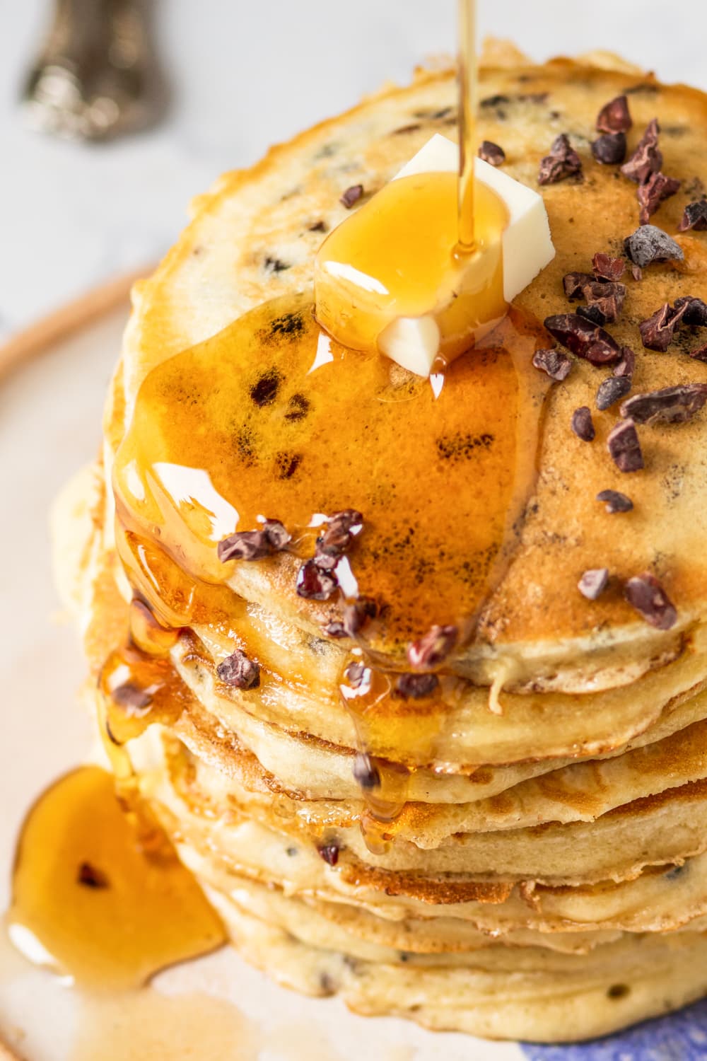 A close up view of a stack of nine chocolate chip pancakes on a white plate with a square slab of butter on the top pancake. Syrup is being poured on the pancakes, dripping down the side of the stack.