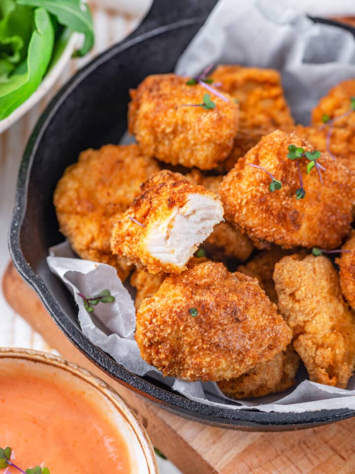 A skillet filled with parchment paper with several keto chicken nuggets on top. One of the chicken nuggets at the front has a bite taken out of it, showing the chicken inside the breading. The skillet is on a wood cutting board on a wooden table. Part of a dipping sauce is in front to the left of the cutting board.