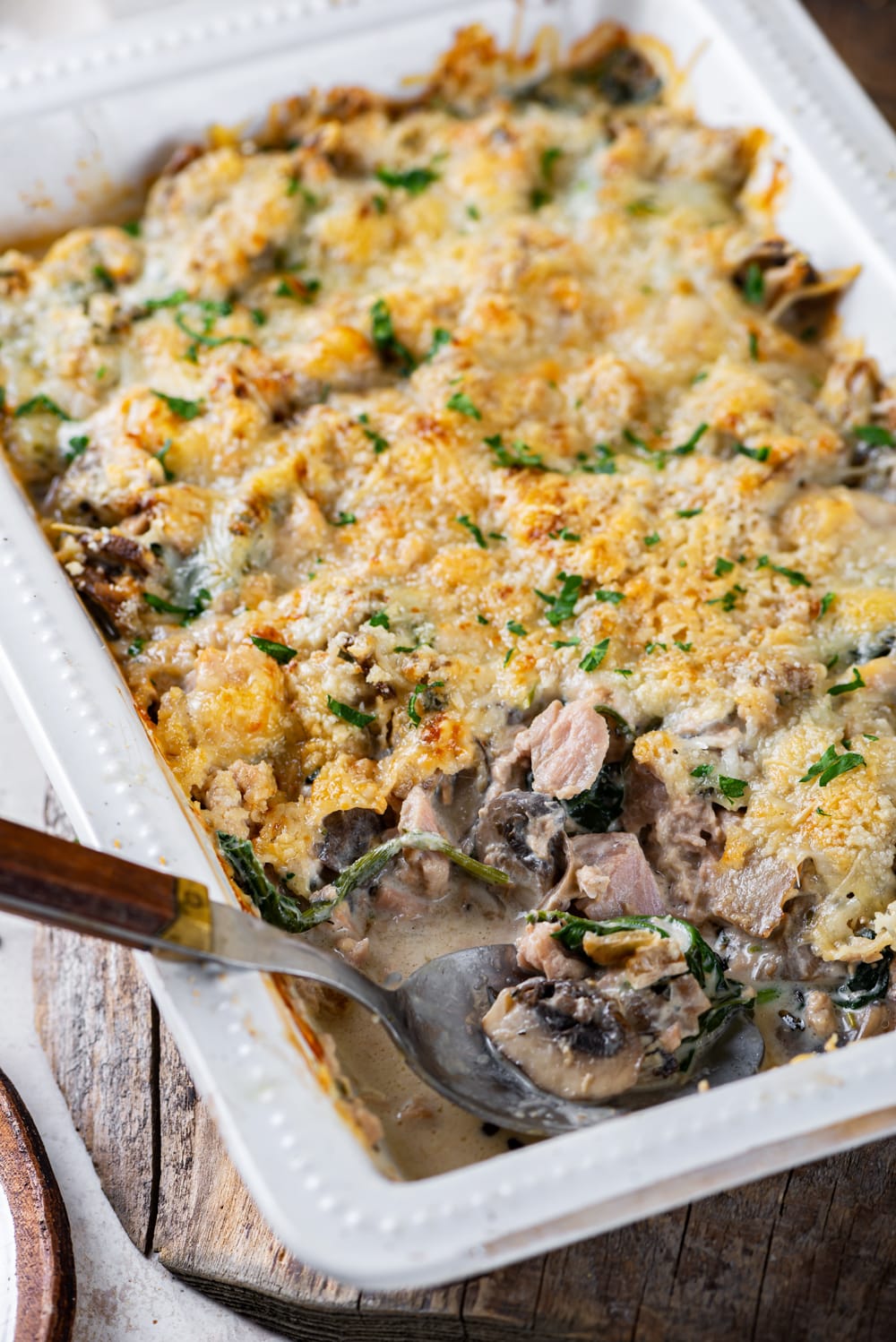 Keto tuna casserole in a white casserole dish. The head of a metal spoon is at the front of the casserole dish and is filled with mushrooms. The casserole dish is on a wooden board.