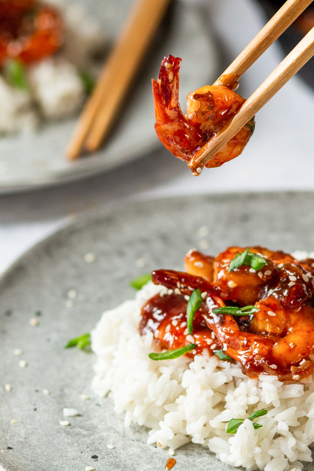 A few pieces of sweet and sour shrimp on a bed of white rice on a grey plate. A pair of chopsticks is holding a piece of shrimp over the shrimp on rice.
