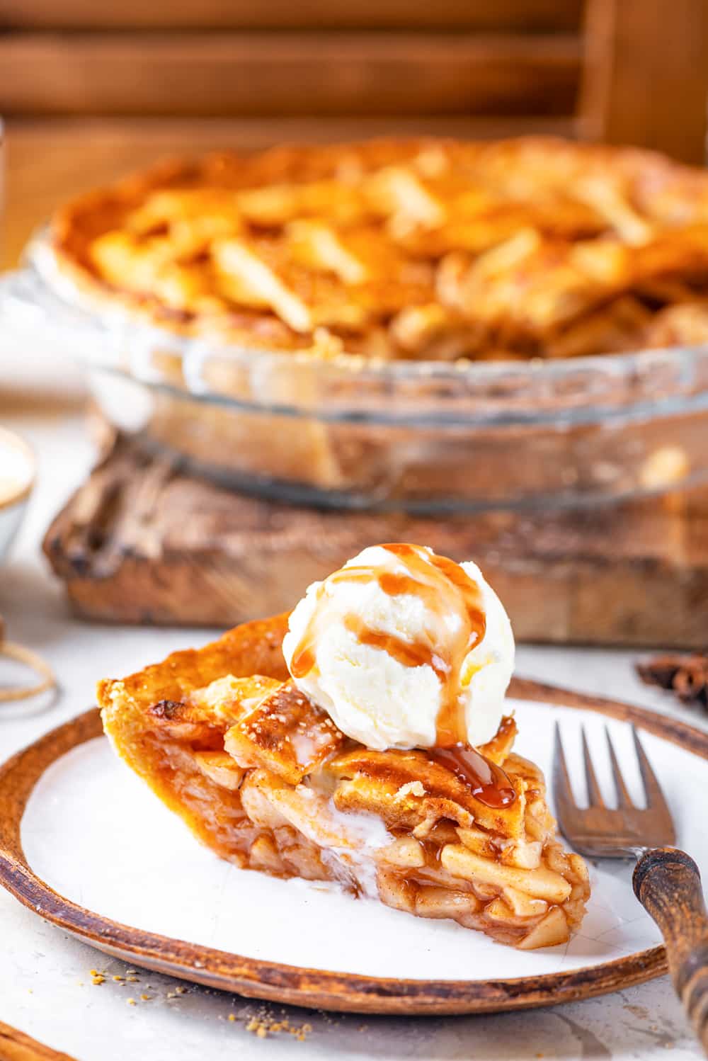 A piece of vegan apple pie with a scoop of vegan vanilla ice cream on top of the piece with a caramel sauce drizzled on the ice cream. There is a fork on the plate to the right of the pie piece. An entire vegan apple pie minus the piece on the plate is behind the plate in glass pie dish on a wooden cutting board. Everything is on a white counter.