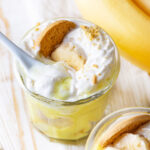 A glass cup filled with vegan banana pudding with vegan whipped cream on top with one slice of a banana and a vanilla cookie in it. There is a white plastic spoon submerged in the top of the whipped cream. Another glass is in front of the other one with only the top visible. The top has a few slices of banana and a vanilla cookie on top of vegan whipped cream. Everything is on a wooden table.