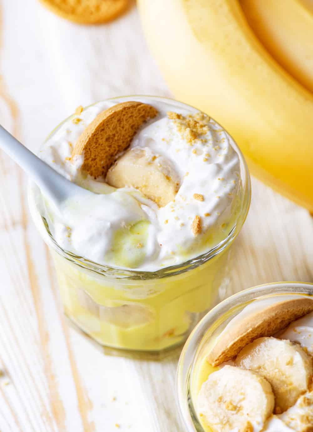 A glass cup filled with vegan banana pudding with vegan whipped cream on top with one slice of a banana and a vanilla cookie in it. There is a white plastic spoon submerged in the top of the whipped cream. Another glass is in front of the other one with only the top visible. The top has a few slices of banana and a vanilla cookie on top of vegan whipped cream. Everything is on a wooden table.