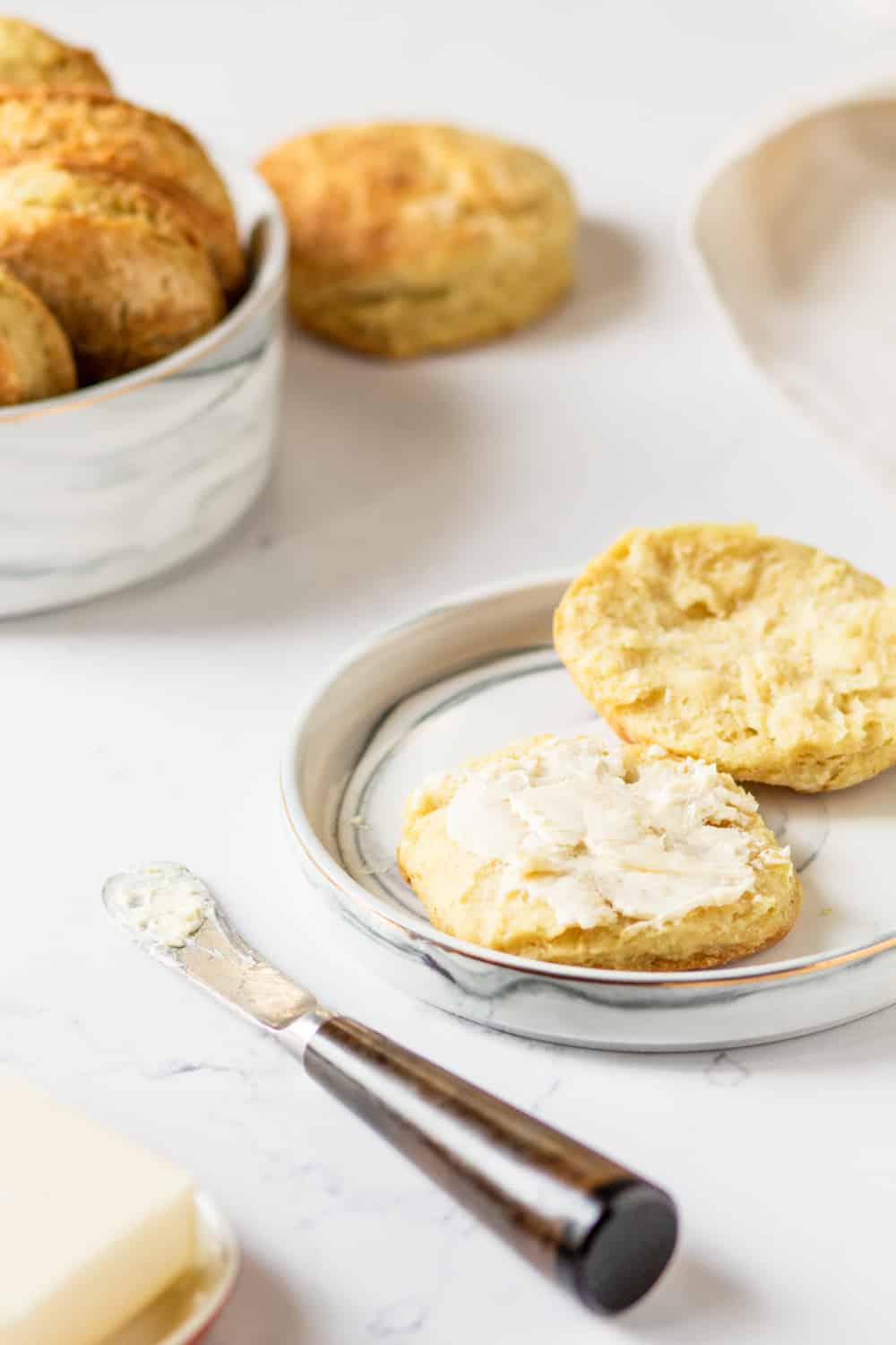 A white plate with a cut open vegan biscuit. The bottom half has a slab of butter on it and is in front of the top half. There is a knife with butter on it in front of the plate and a stick of butter in front of the knife. There is a white bowl filled with vegan biscuits behind plate to the left. A whole vegan biscuit is on the right side of the bowl. Everything is on a white counter.