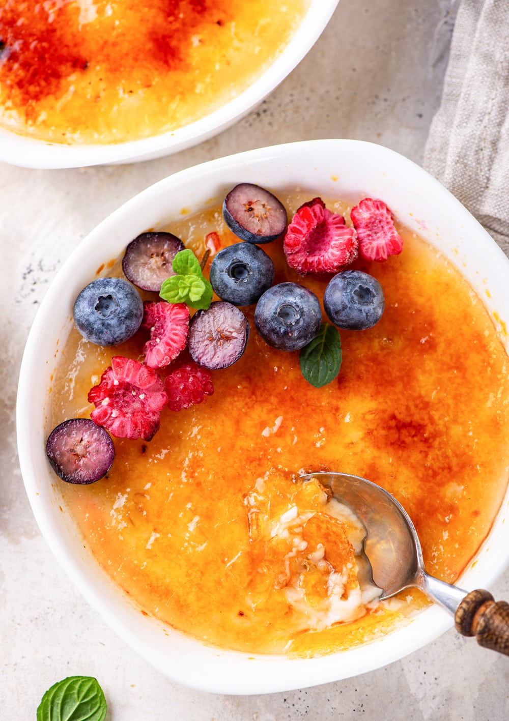 An overhead view of vegan creme brulee in a ramekin. There are sliced raspberries and blueberries at the back half of the vegan creme brulee. A spoon is scooping in to the vegan creme brulee at the front of the ramekin. The ramekin is set on a white counter.
