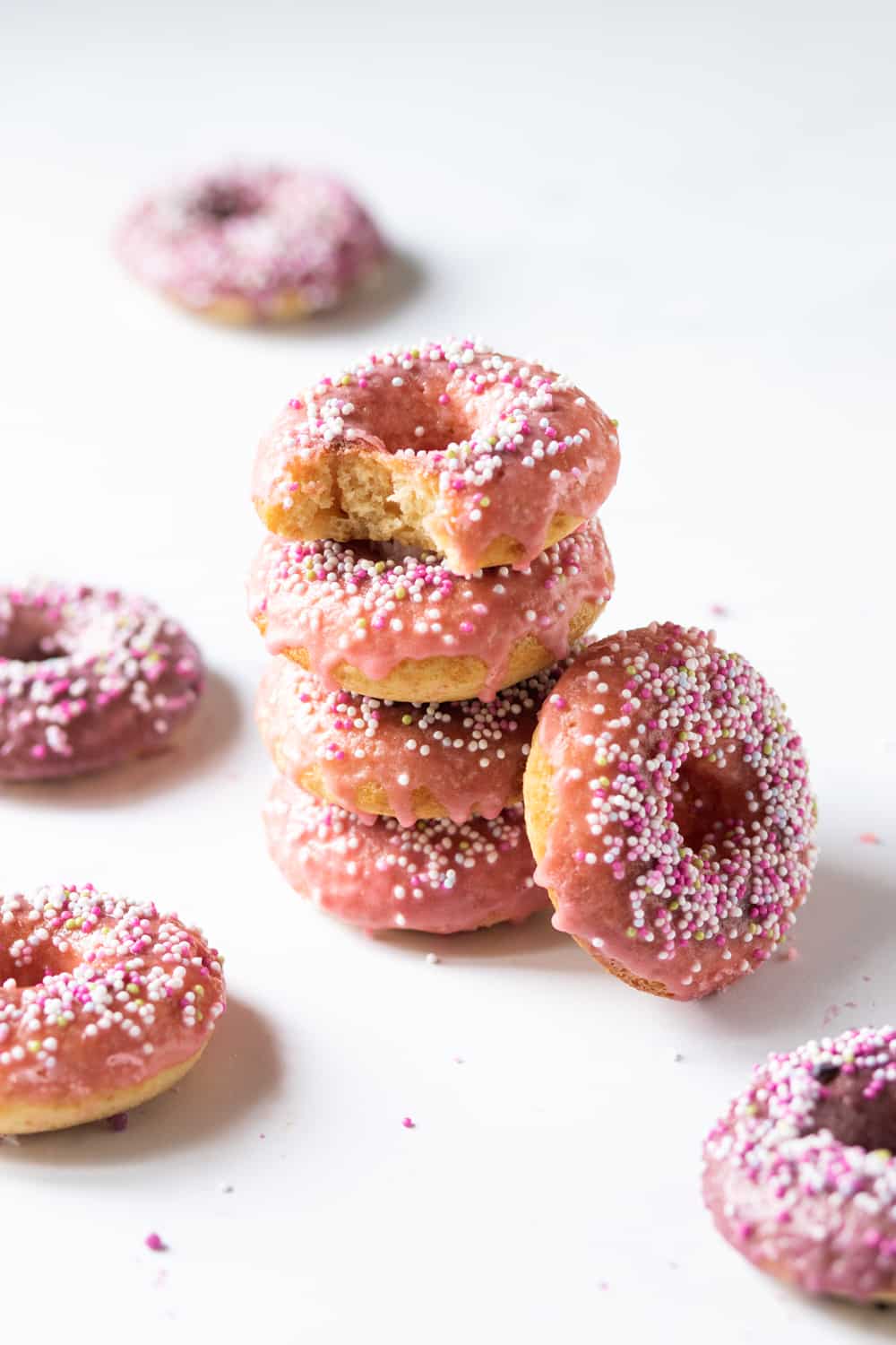A stack of four vegan donuts with pink glaze and white, pink, and yellow ball sprinkles on top. The donut of top of the stack has a bite taken out of it. There is a whole donut on its side leaning agains the stack. One donut is laying flat in front of it, another donut is in front of the stack, another to the left of it, and one behind it. All the donuts are on a white counter.