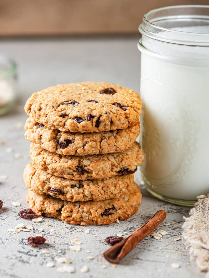 Five vegan oatmeal raisin cookies stacked on top of one another. A tall glass jar of milk is to the right of the cookies. A cinnamon stick is in front of the cookies and milk with a few raisins and oats scattered around them. Everything is on a grey table.