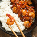 An overhead view of a skillet half filled with white rice on the left and half filled with firecracker shrimp on the right. One shrimp is being held between a pair of chopsticks over the rice.