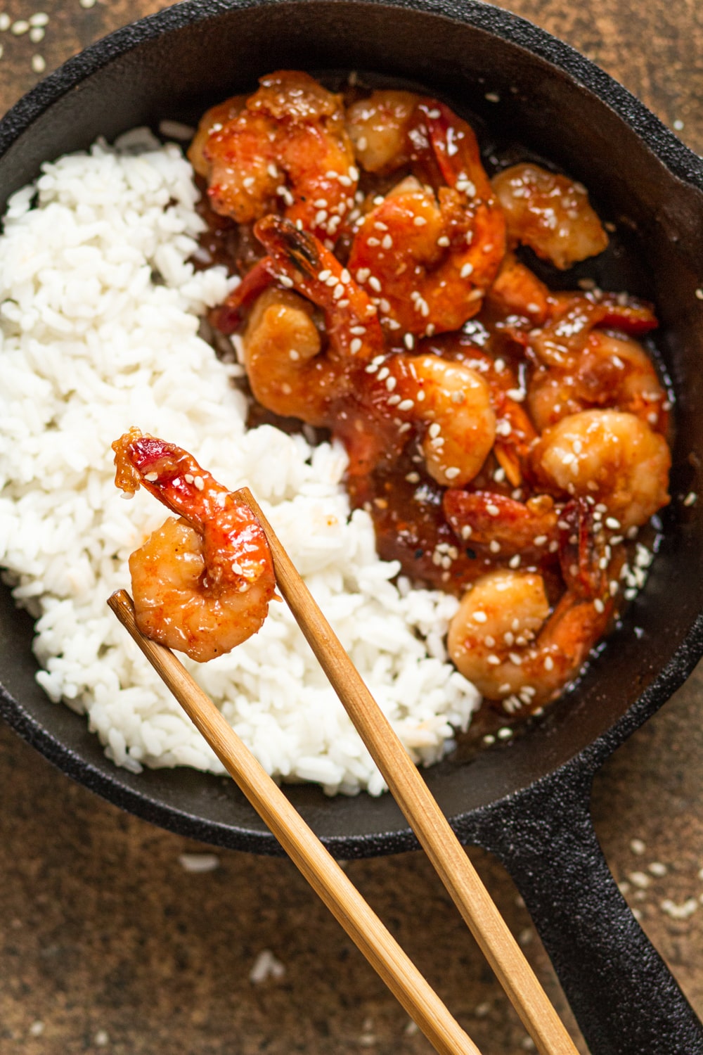 An overhead view of a skillet half filled with white rice on the left and half filled with firecracker shrimp on the right. One shrimp is being held between a pair of chopsticks over the rice.