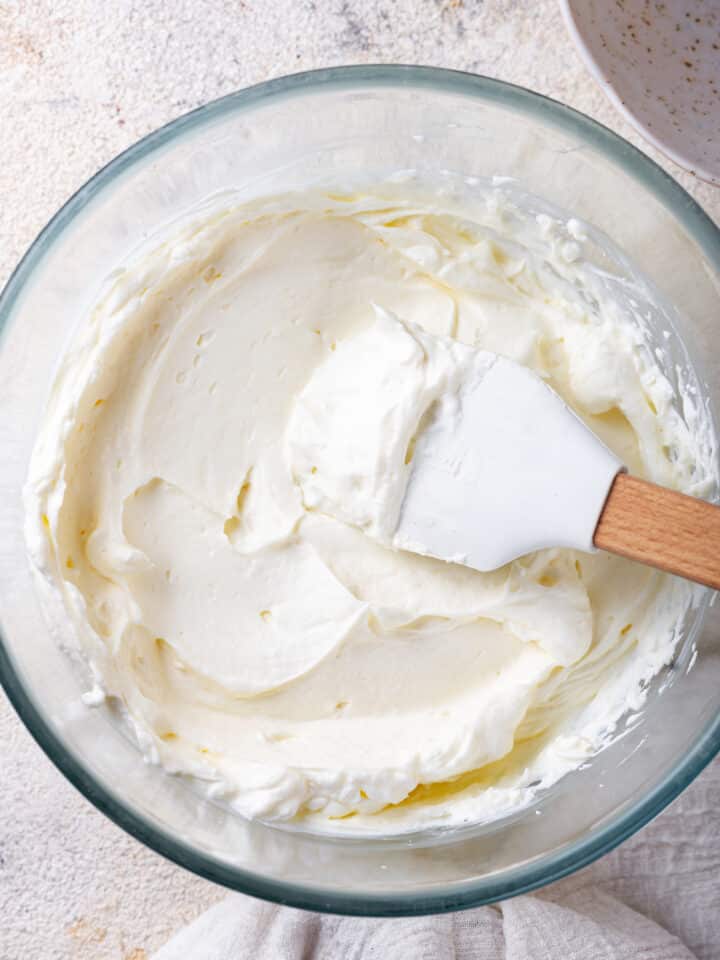 An overhead view of a glass bowl with keto cream cheese frosting in it. A rubber spatula is in the bowl with the wooden shaft leaning against the right side of the bowl and the spatula head just above the frosting with frosting on it.