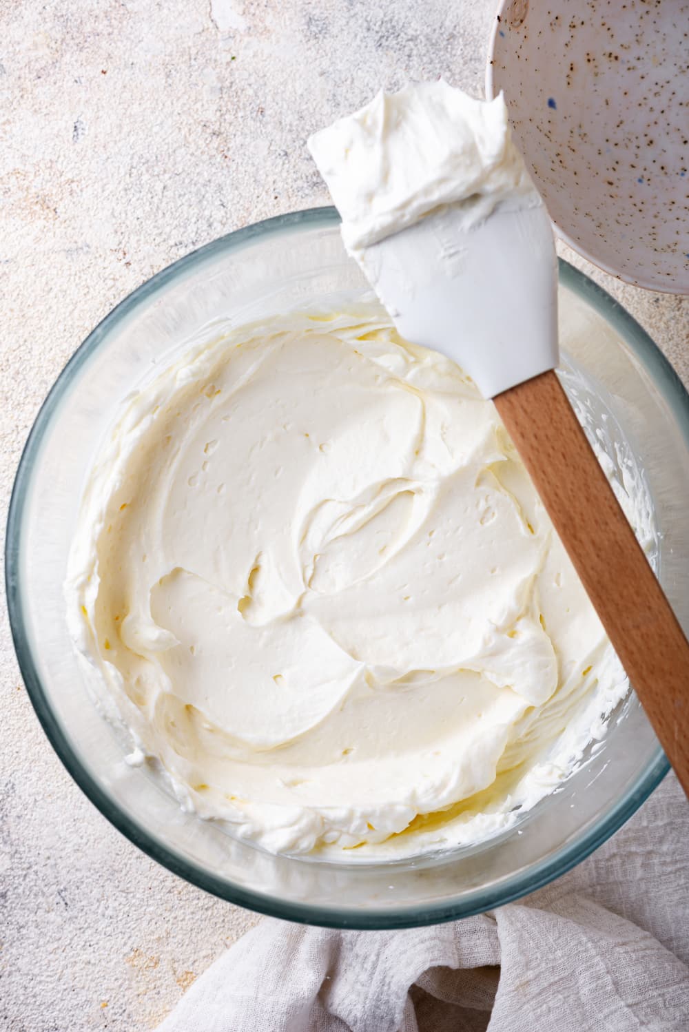 An overhead view of a glass bowl that is filled with keto cream cheese frosting. A rubber spatula is hovering over the right side of the bowl and there is frosting on the spatula head.