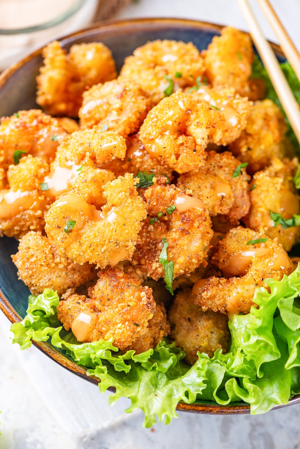 Bang bang shrimp in a blue bowl with a lettuce garnish at the front of the bowl. There is a set of chopsticks laying across the right rim of the bowl. The bowl is on a white counter top.