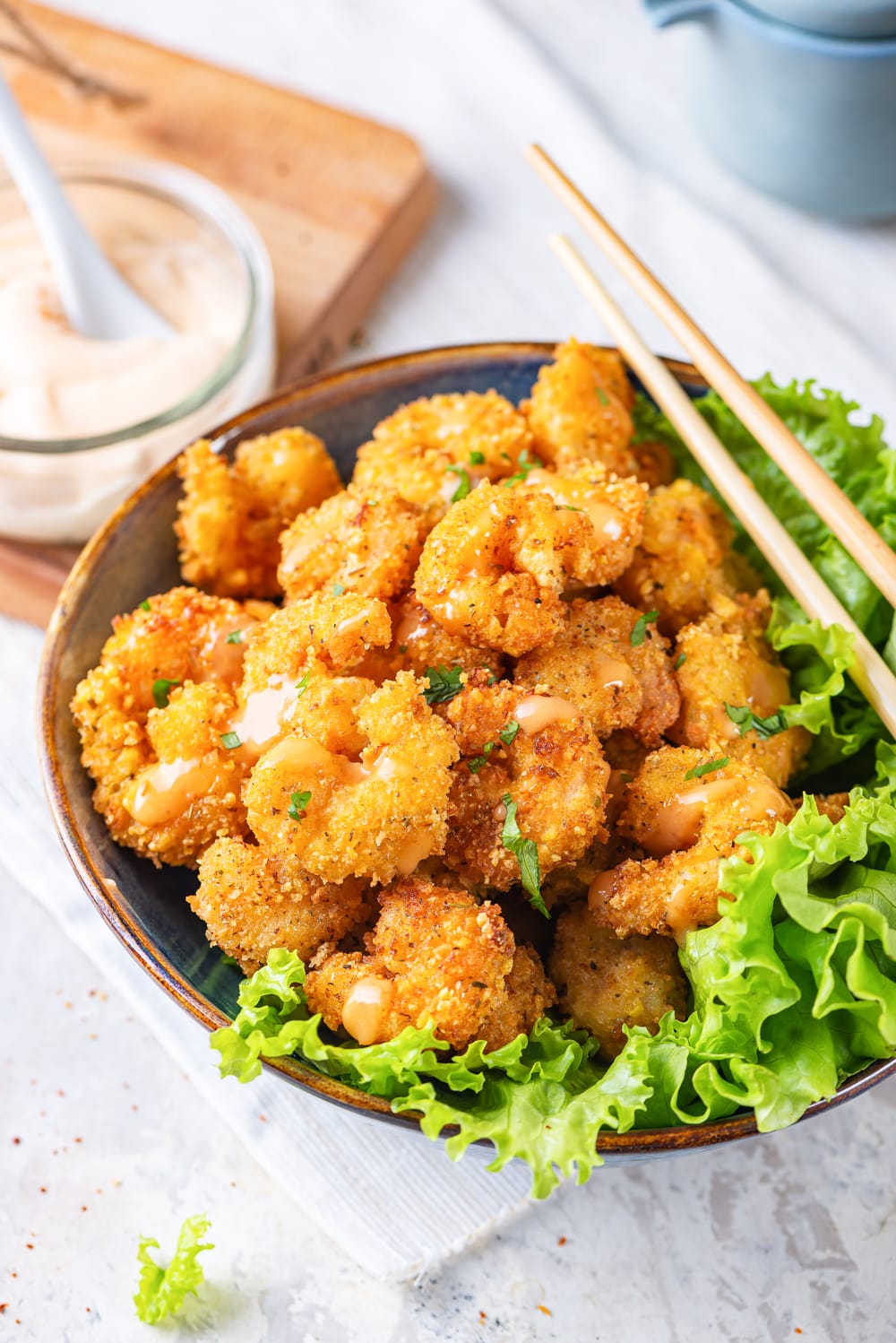 A blue bowl filled with bang bang shrimp, with a big leaf of lettuce on the rim of the right half of the bowl. A glass jar of bang bag sauce is on a wooden cutting board to the back left of the bowl. Everything is on a white table cloth that is on a white counter.