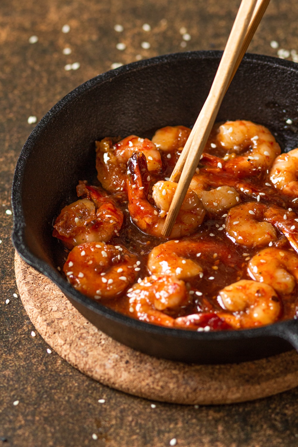 Several pieces of shrimp in a firecracker sauce in a skillet. There is a set of chopsticks grabbing a piece of shrimp from the skillet. The skillet is on a circular cork board which is on a brown counter.