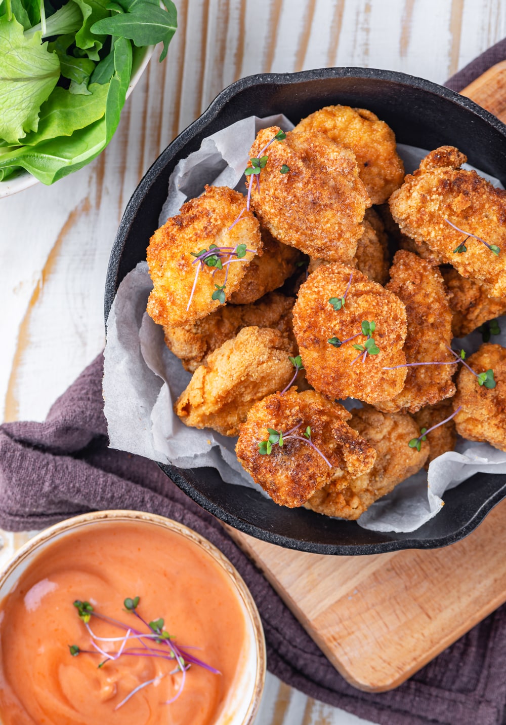 An overhead view of keto chicken nuggets on top of parchment paper in a skillet on a wood cutting board. In front of the cutting board is dipping sauce in a bowl and everything is on a wooden table.