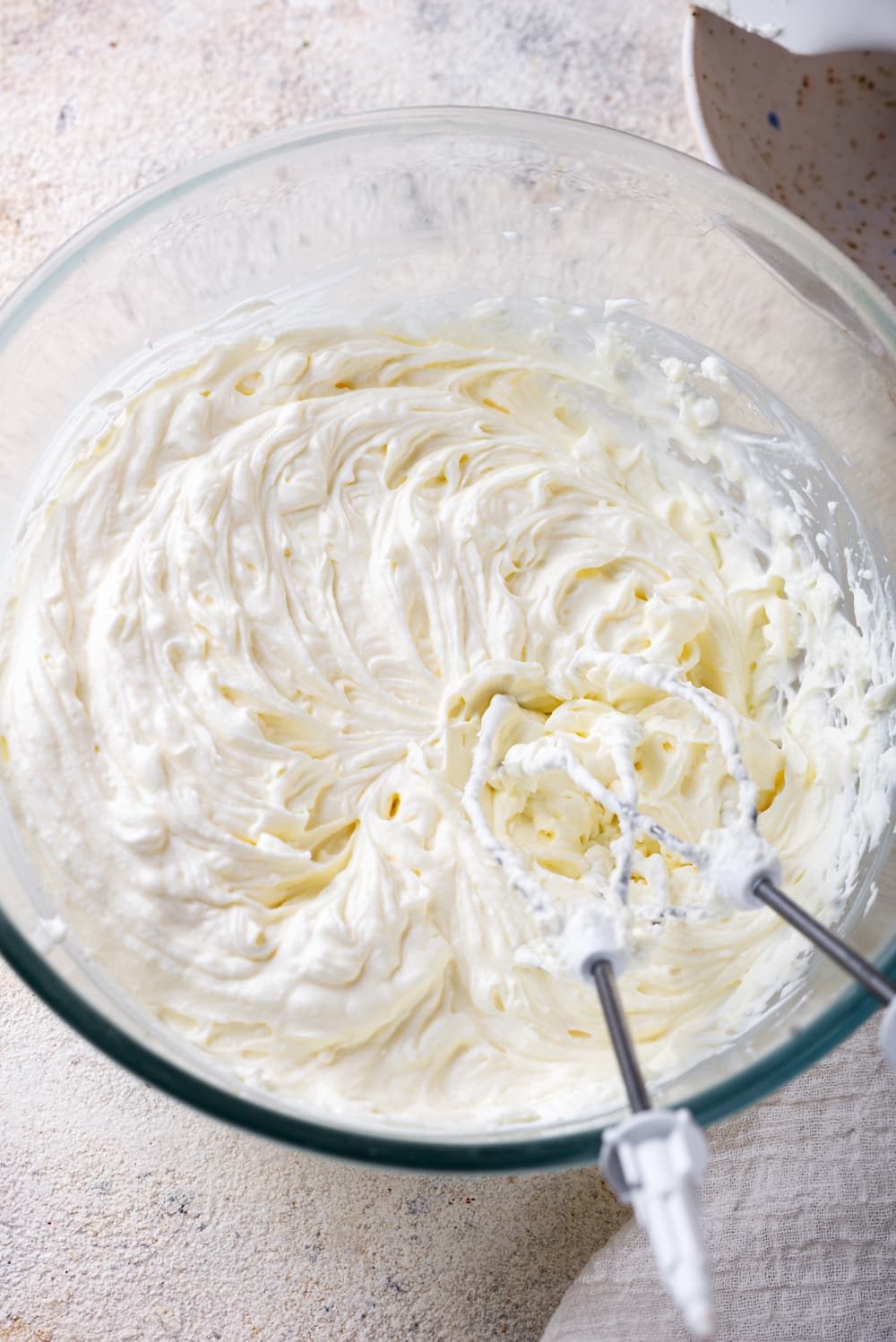 A glass bowl of whipped keto cream cheese frosting. The head of the mixers are in the frosting and the stems are leaning against the front side of the bowl.