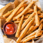 An overhead view of a basket of keto french fries. The french fries are laying on top of a sheet of brown newspaper. A small cup of ketchup is on the left side of the fries in the basket. The basket is on a white counter.