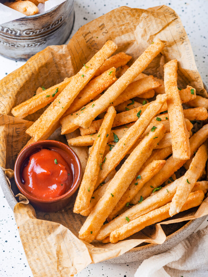 An overhead view of a basket of keto french fries. The french fries are laying on top of a sheet of brown newspaper. A small cup of ketchup is on the left side of the fries in the basket. The basket is on a white counter.