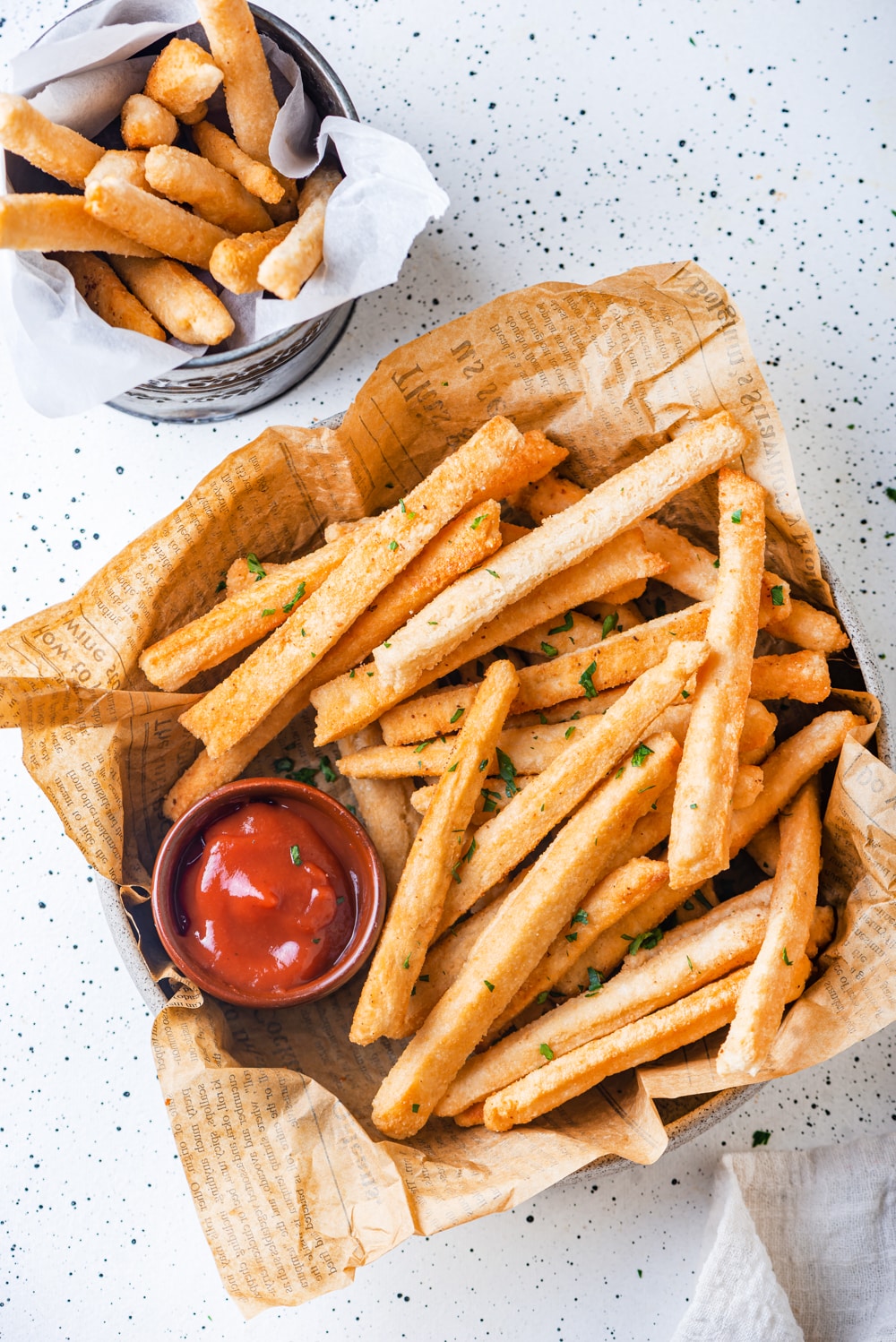 An overhead view of a basket lined with brown newspaper and filled with keto french fries. There is a small cup of ketchup placed in the left of the basket. A metal cup filled with parchment paper and keto french fries on top is behind the basket. The basket and cup are on a white table.