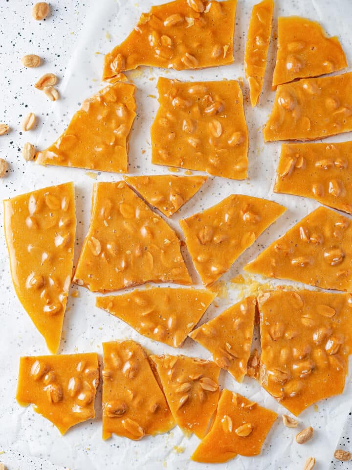 An overhead view of small and large pieces of peanut brittle scattered on a sheet of parchment paper. There are peanuts around the sheet of parchment paper and everything is on a white counter.