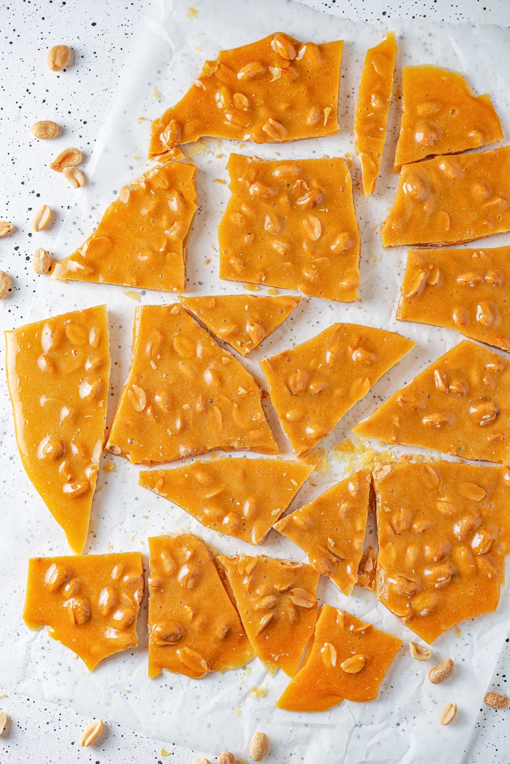 An overhead view of small and large pieces of peanut brittle scattered on a sheet of parchment paper. There are peanuts around the sheet of parchment paper and everything is on a white counter.