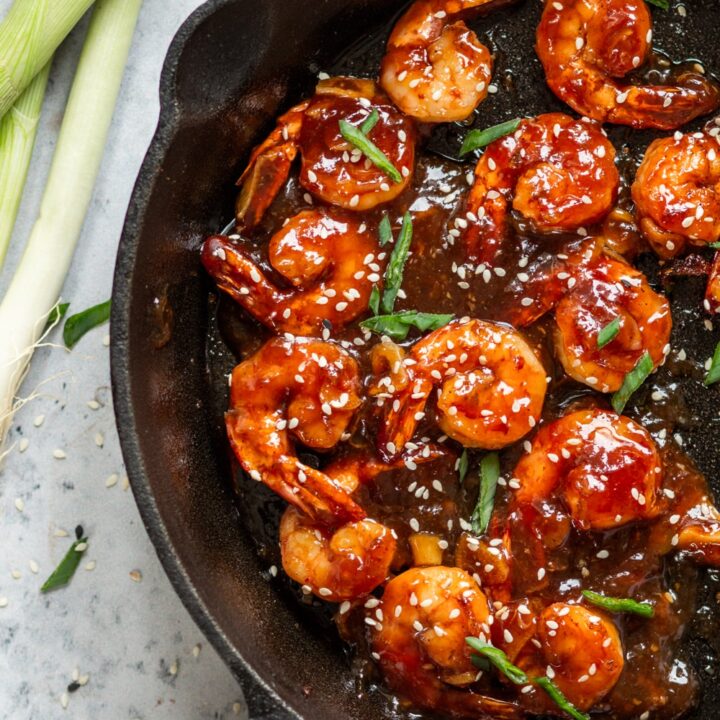 An overhead view of several pieces of shrimp covered in a sweet and sour sauce in a skillet. There are sesame seeds and sliced green onion on top of the shrimp. A few stalks of green onion are to the left of the skillet and everything is on a white counter.