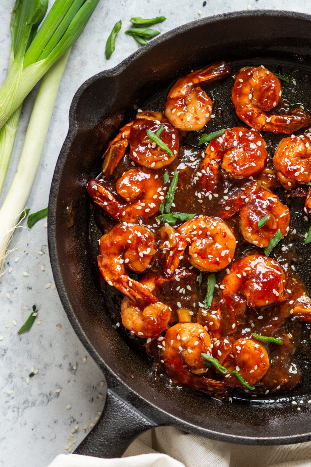 An overhead view of several pieces of shrimp covered in a sweet and sour sauce in a skillet. There are sesame seeds and sliced green onion on top of the shrimp. A few stalks of green onion are to the left of the skillet and everything is on a white counter.