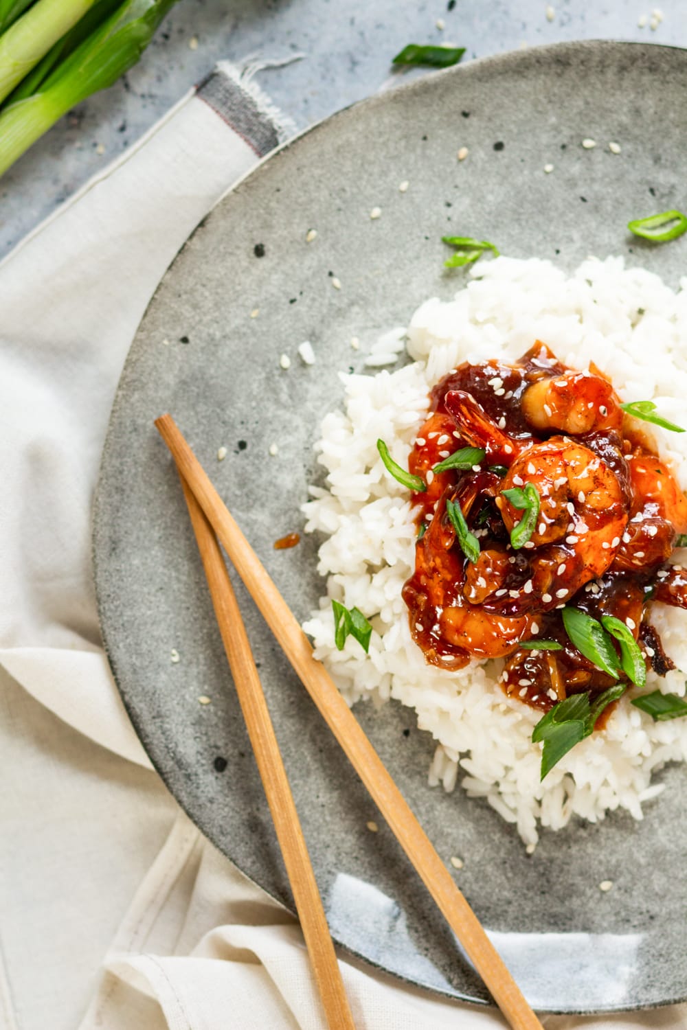 An overhead view of a few pieces of sweet and sour shrimp on a pile of white rice on a grey plate. There are sesame seeds and sliced green onions on the shrimp. A pair of chopsticks is on the plate next to the left of the shrimp. The plate is on a bunched white tablecloth.