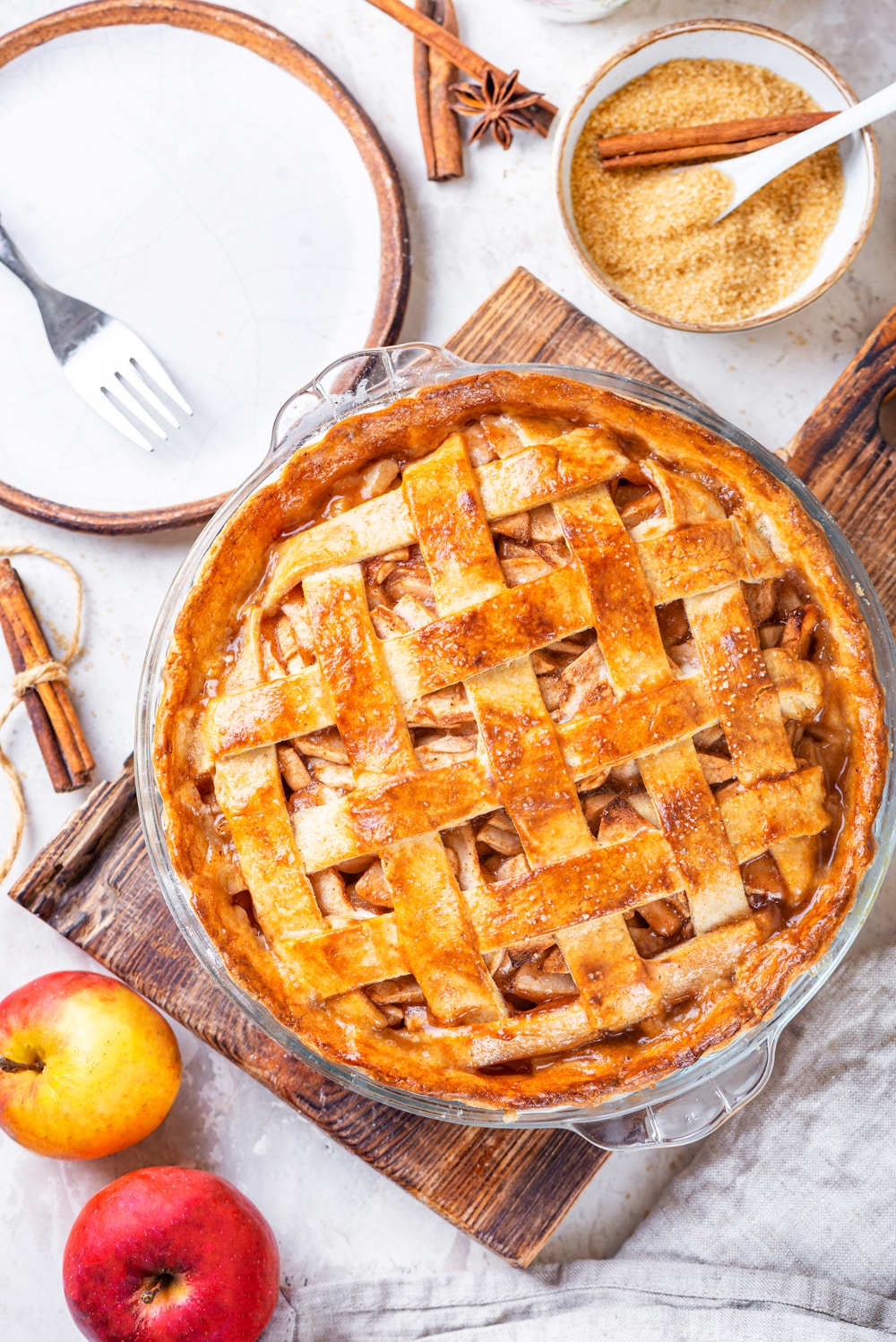 An overhead view of a vegan apple pie in a glass pie dish on a wooden cutting board. There are two apple in front of and to the left of the cutting board. A white plate with a fork on it and a white bowl filled with sugar is behind the cutting board. All of this is on a grey cutting board.