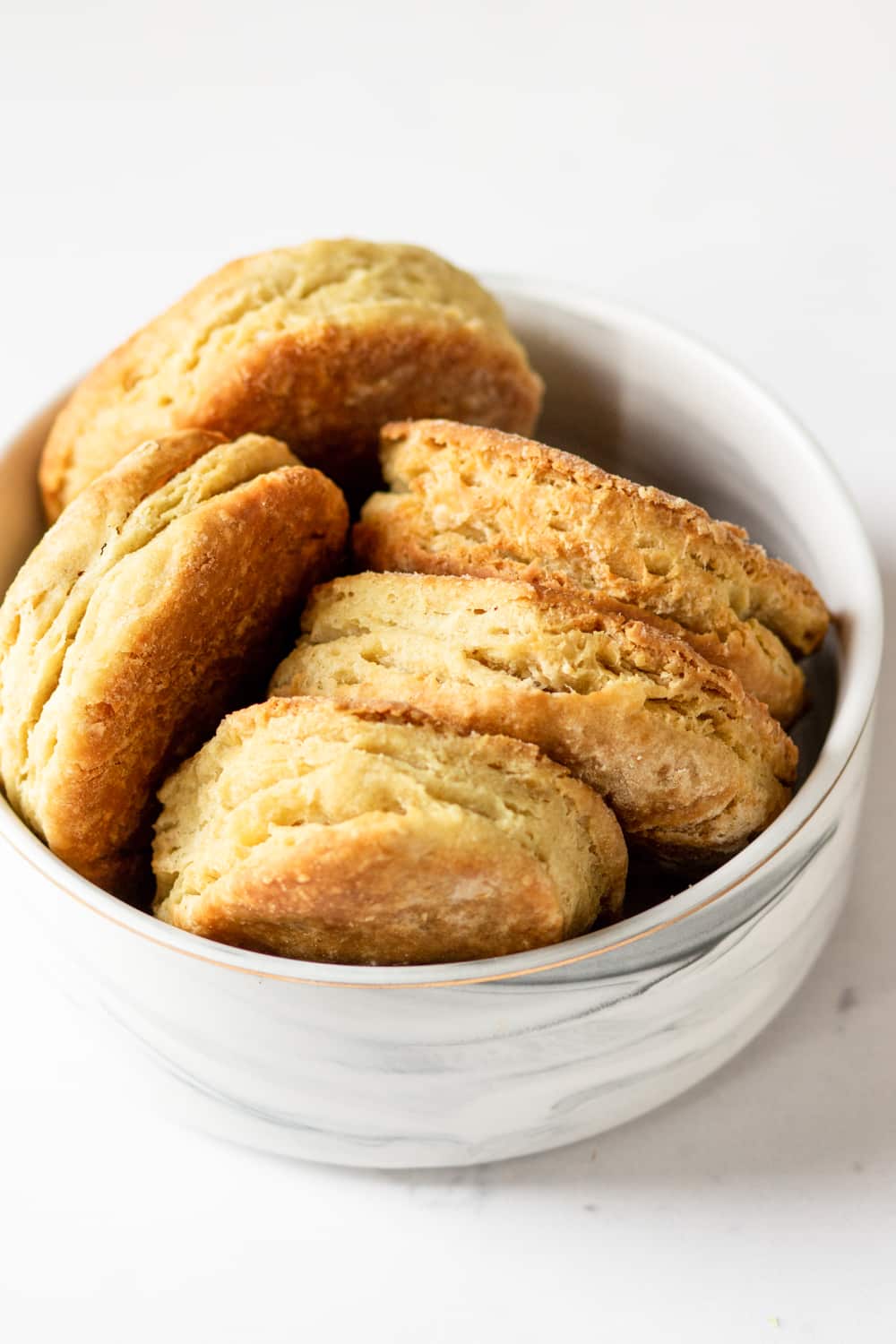 A white bowl filled with five vegan biscuits. Three of them are in a row and two are to the left and all of the biscuits are on their side. The bowl is on a white counter.