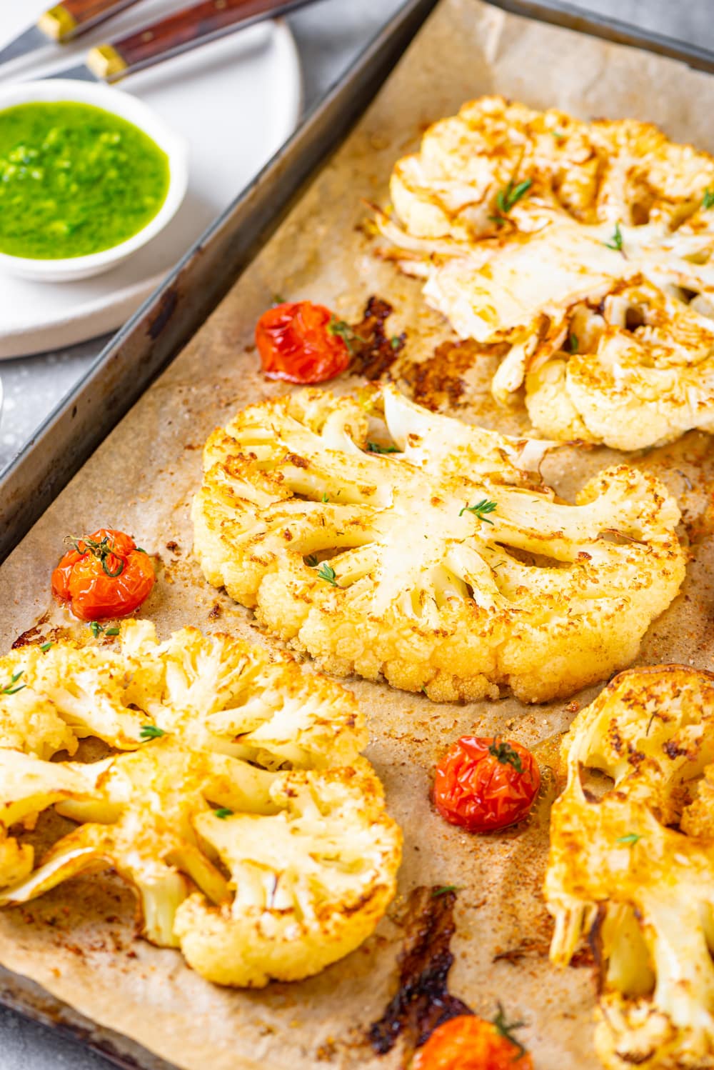 A close up view of three cauliflower steaks lined up on a baking sheet lined with parchment paper. There is three cherry tomatoes on the baking sheet around the cauliflower. Behind the baking sheet to the left isa white plate with a small white bowl of cilantro sauce on it.