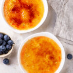An overhead view of two ramekins filled with vegan creme brulee. The ramekins are on a white counter with one ramekin in front of the other. There is a small bowl of blueberries off to the left, in the middle of the ramekins. A spoon is in front of the front ramekin.