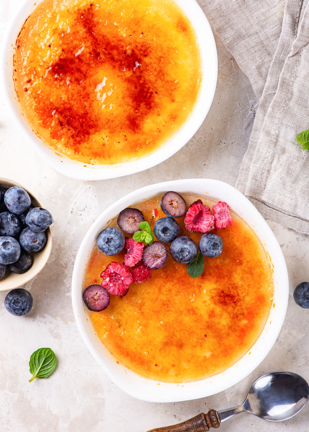 An overhed view of two ramekins filled with creme brulee with one ramekin in front of the other. The ramekin in the front has sliced raspberries and blueberries on the back half of the vegan creme brulee. A small bowl of blueberries is to the left, between the ramekins. A spoon is front of the first ramekin and everything is on a white counter.