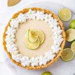An overhead view of a whole vegan key lime pie. The pie is on a circular white marble board with limes at the right edge of it. The marble board is on a white counter.