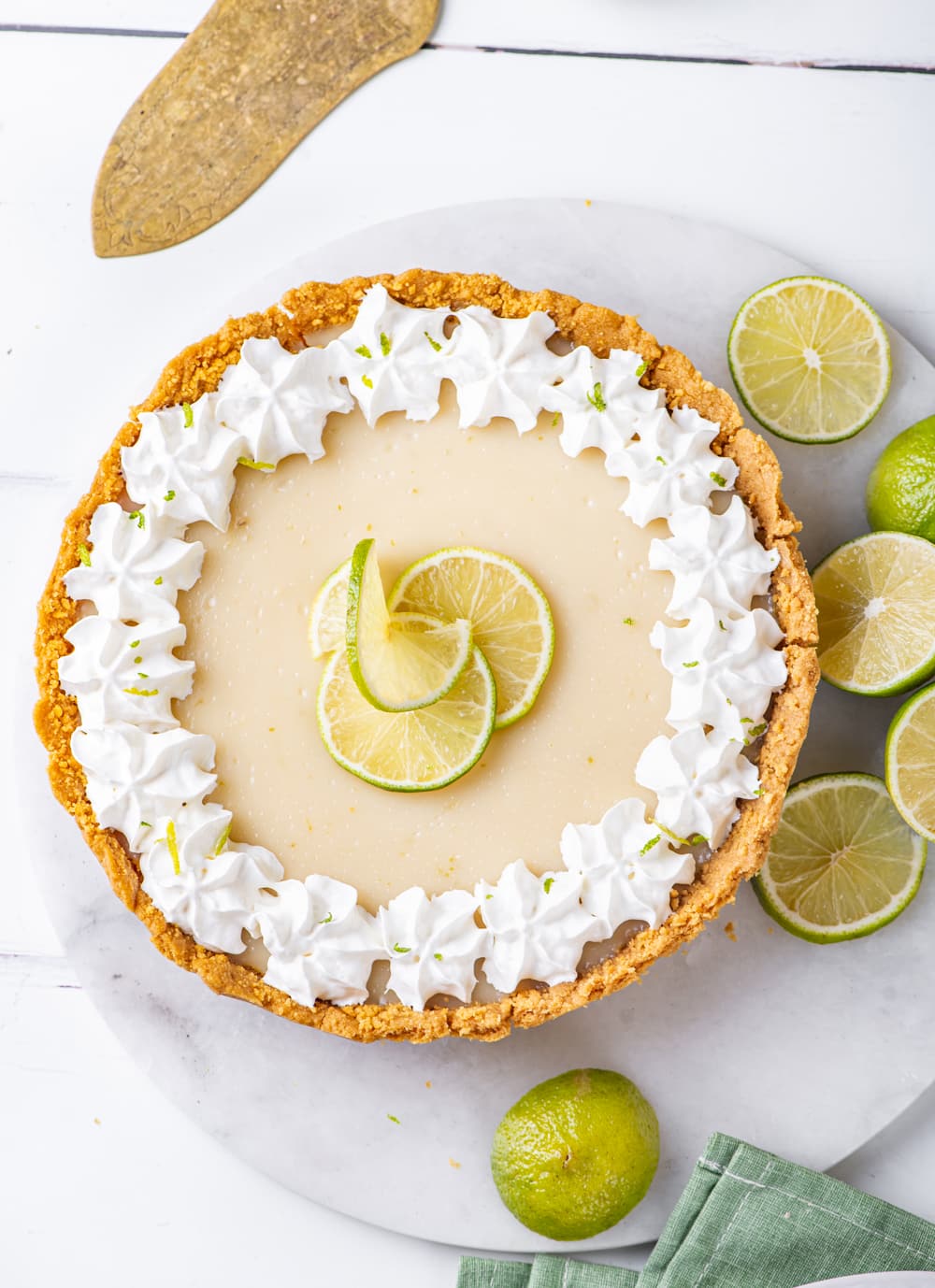 An overhead view of a whole vegan key lime pie. The pie is on a circular white marble board with limes at the right edge of it. The marble board is on a white counter.